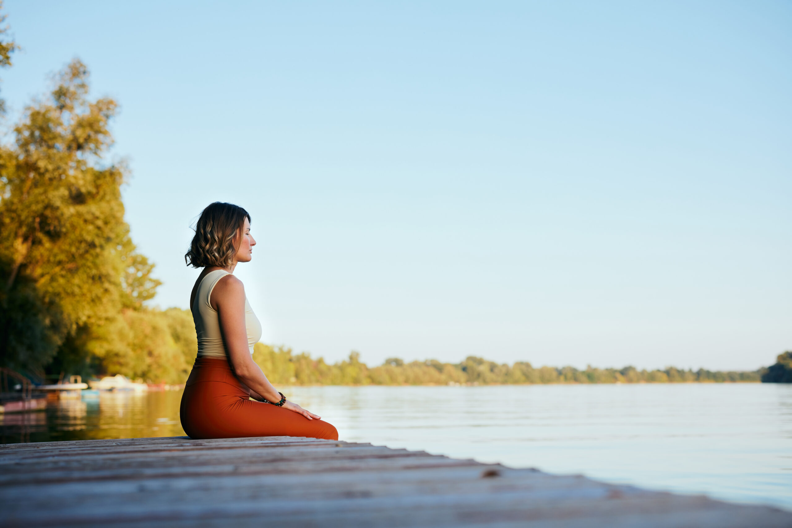 A middle-aged woman is relaxing on the dock and doing yoga breathing exercises. She put her feet in the water.
